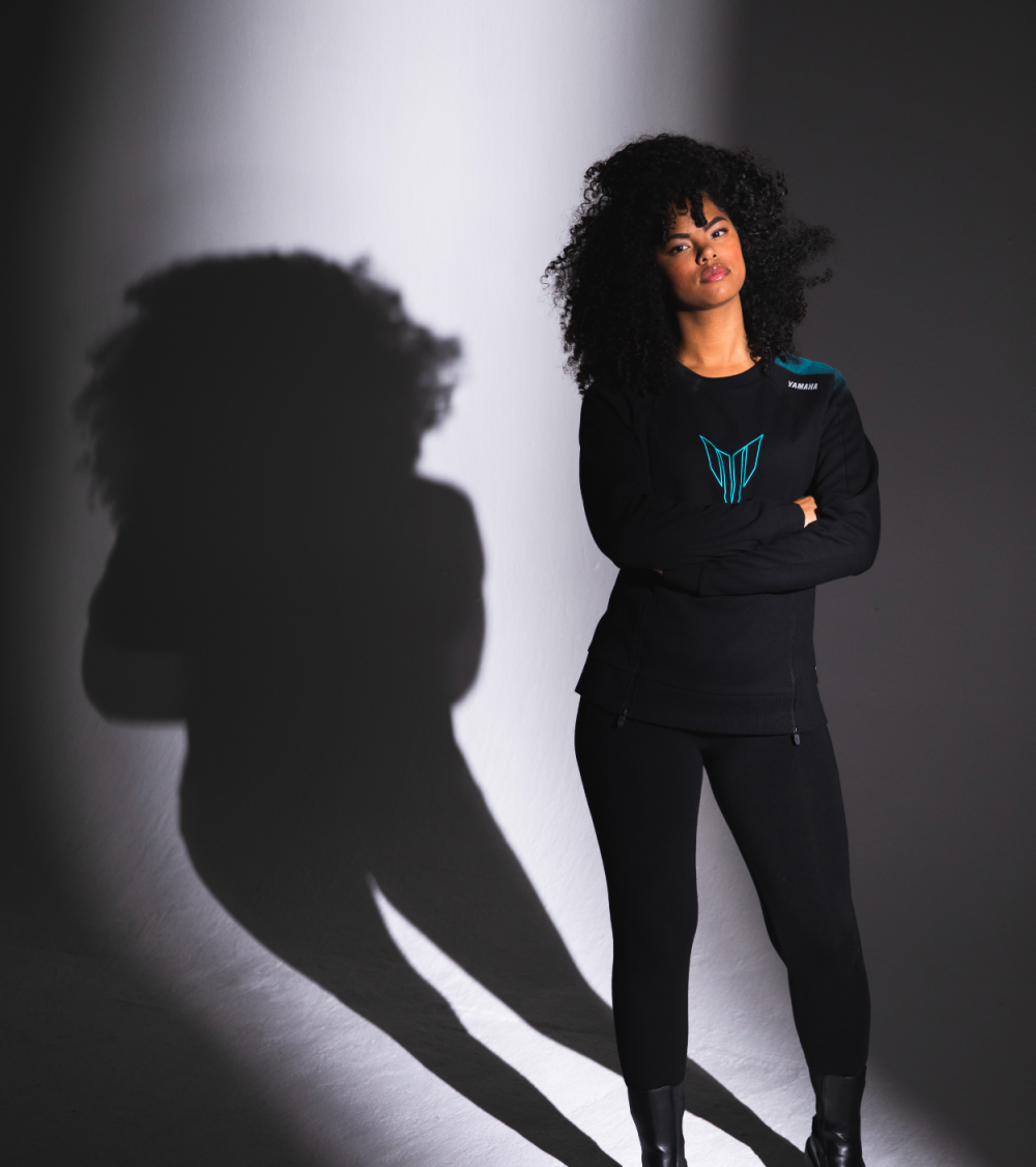 Black woman in Yamaha clothes with blue colors, looking straight into the camera, lit up with a spotlight.