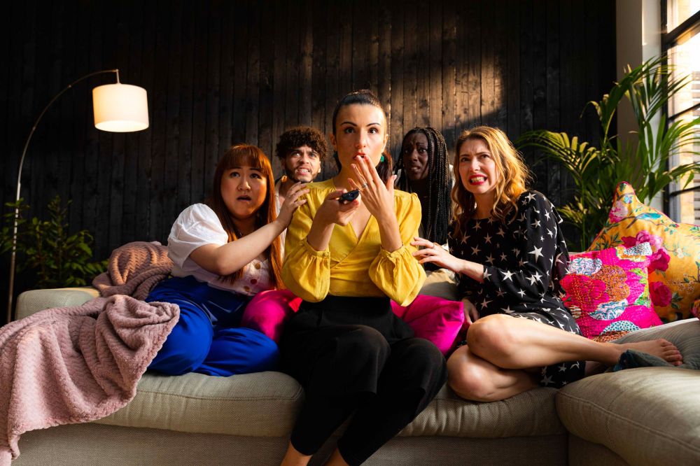 A group of people on the sofa watching a television in suspense in the early evening. The group is young but diverse.