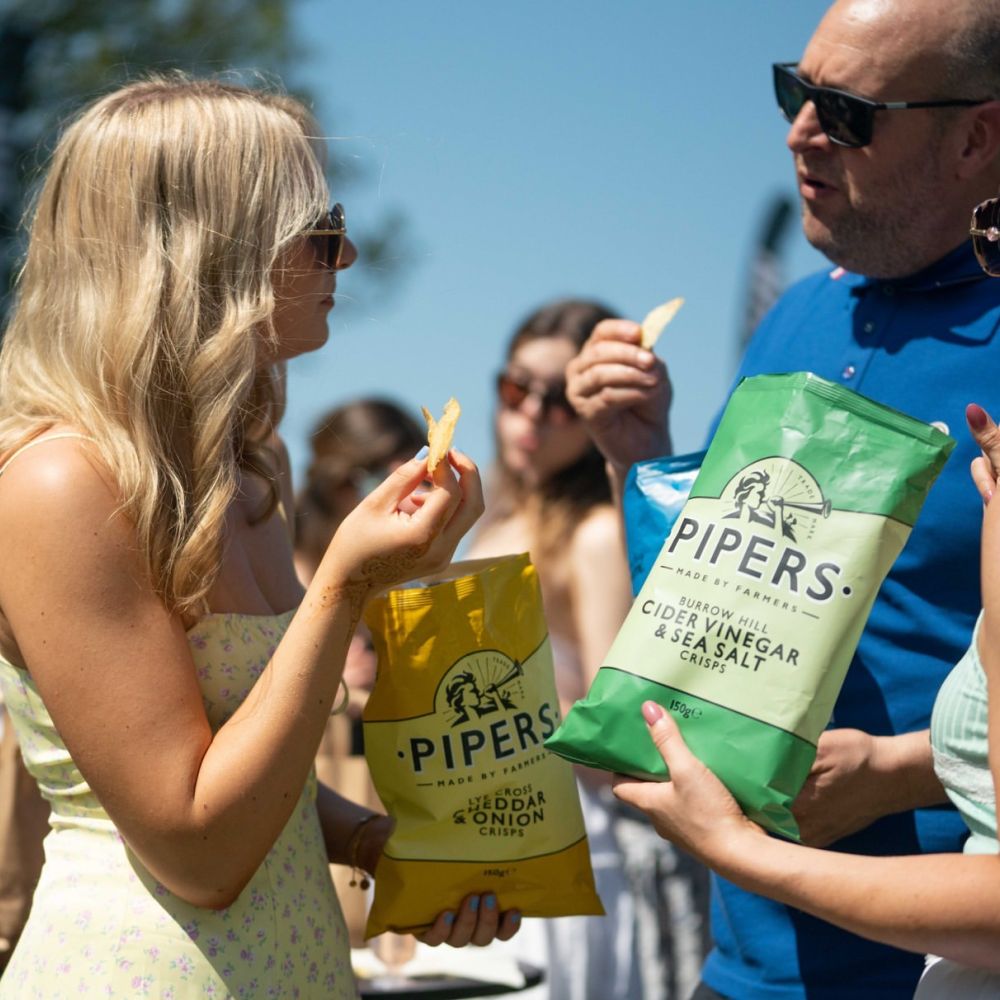 People eating large bags of Pipers crisps in the Sun with sunglasses on.