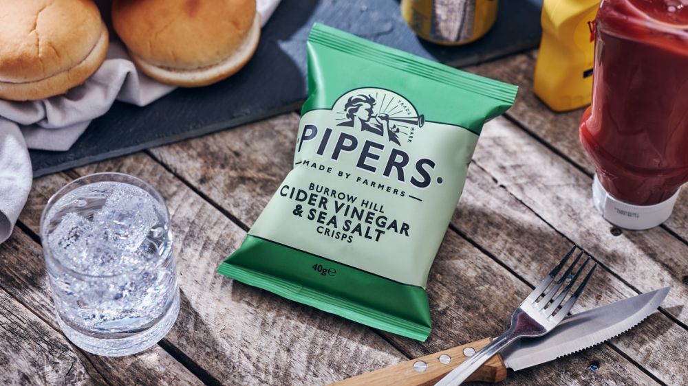 A green packet of Pipers Cider Vinegar & Sea Salt Crisps on a picnic-style table with ketchup, buns and a knife and fork.