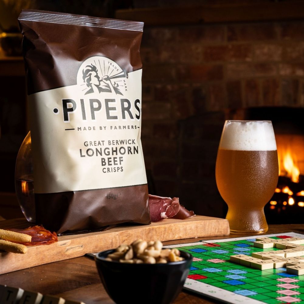 Pipers Great Berwick Longhorn Beef Crisps shown on a table with beer, scrabble, and peanuts.