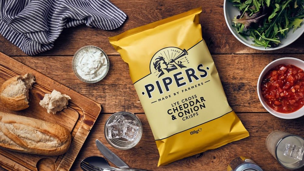 Overhead shot of a yellow packet of Pipers Lye Cross Cheddar & Onion Crisps on a picnic-style table including salsa, salad bread and a teatowel.