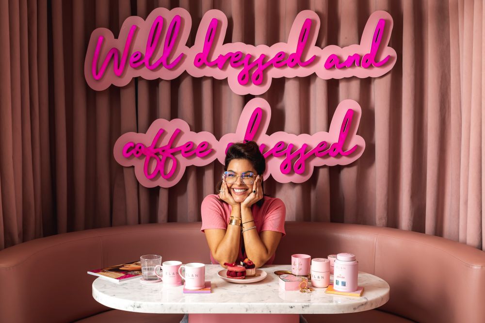 Woman on a rouge leather sofa in an El & N store with pink curtains behind her. The woman is leaning her elbows on a marble table and smiling.