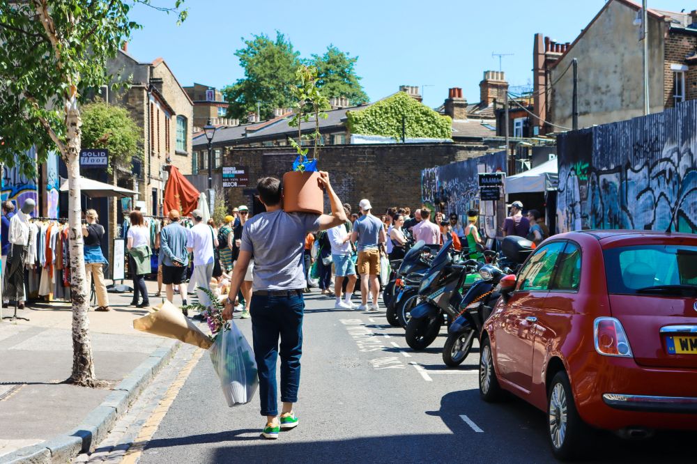 A man carrying a flower pot on his shoulder walking down Columbia Road Flower market. It's a sunny day and there are mopeds and cars parked on the side of a narrow street.