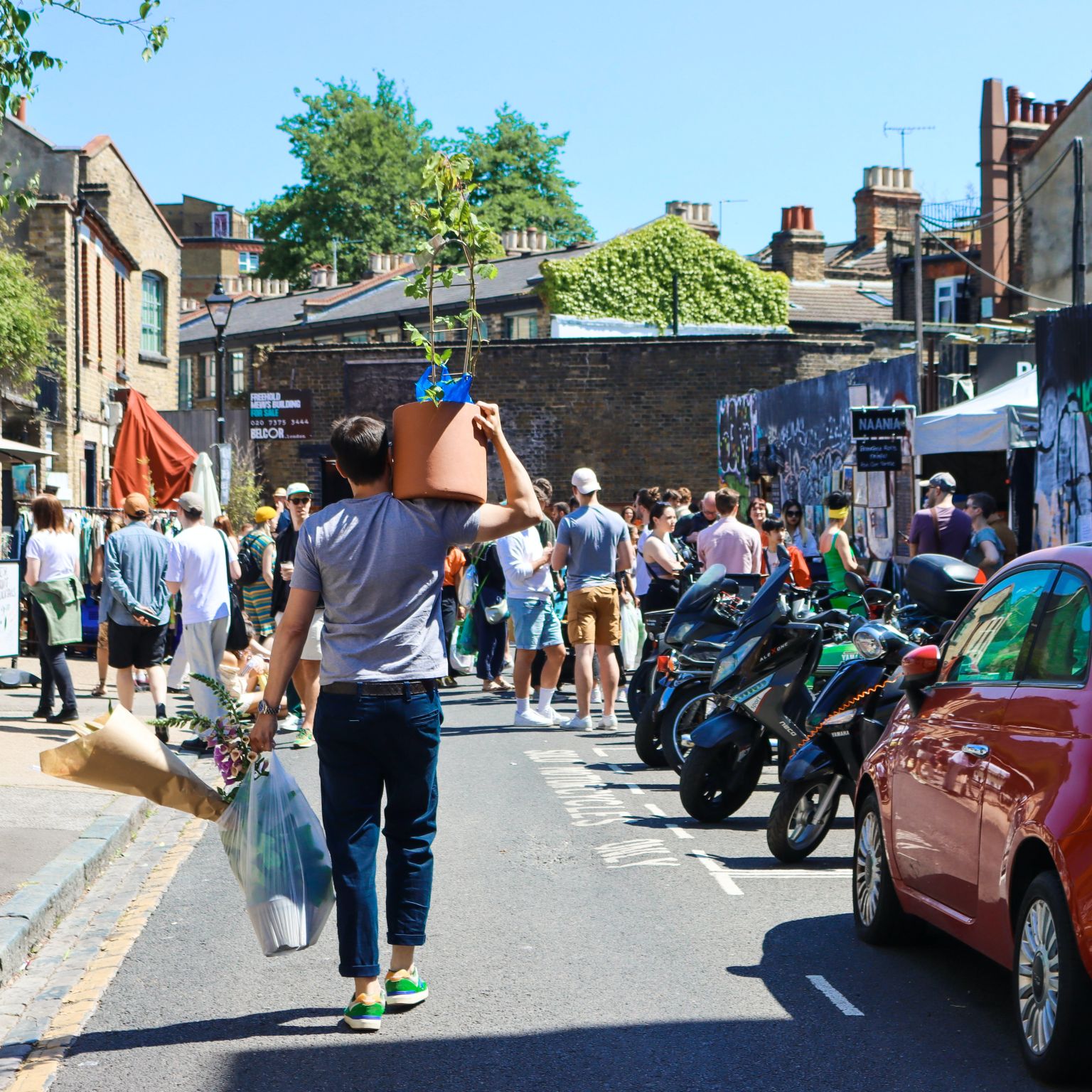 A man carrying a flower pot on his shoulder walking down Columbia Road Flower market. It's a sunny day and there are mopeds and cars parked on the side of a narrow street.