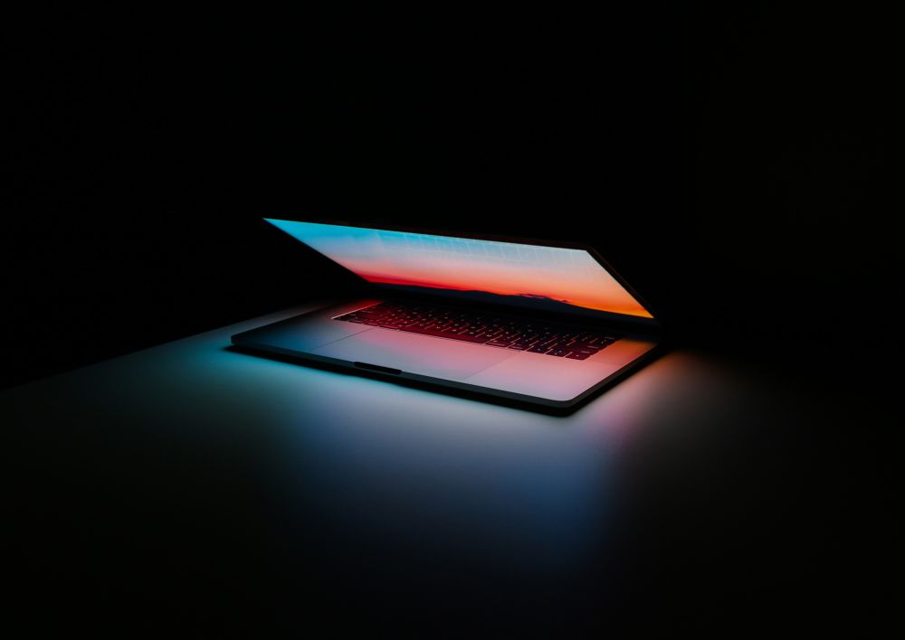 A laptop lid opening with a sunset screen wallpaper in a dark room.