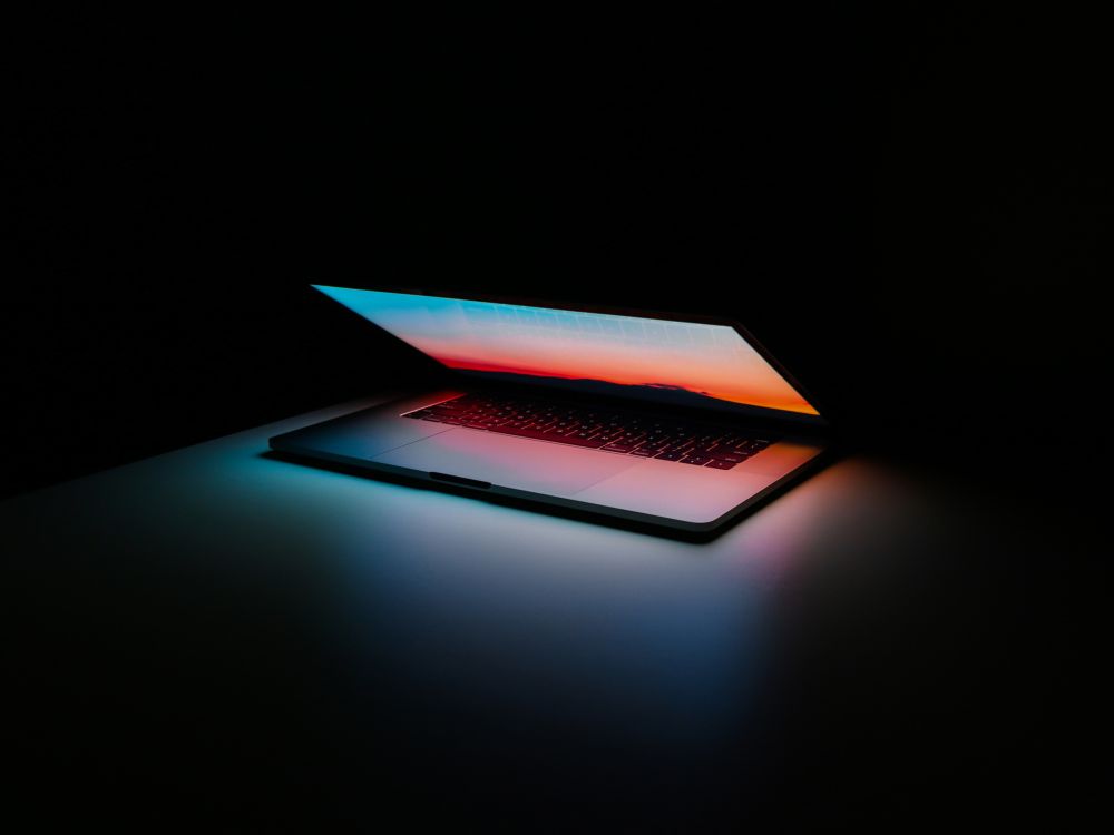 A laptop lid opening with a sunset screen wallpaper in a dark room.