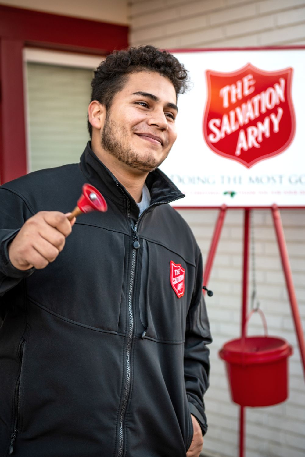A young latino looking man rings a bell smiling, with a Salvation Army placard in the background.