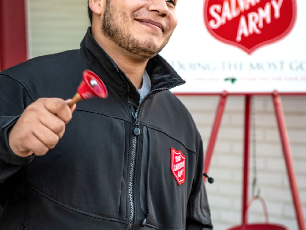 A young latino looking man rings a bell smiling, with a Salvation Army placard in the background.