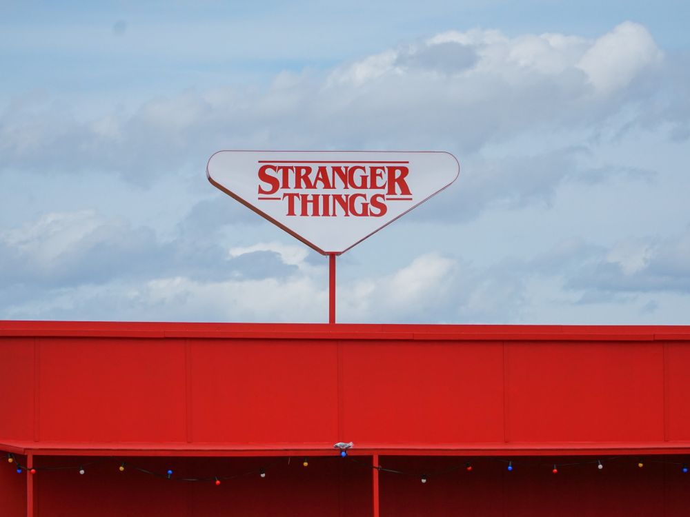 A triangular Stranger Things sign above a red building. There are clouds in the background behind the building.