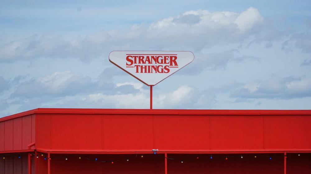 A triangular Stranger Things sign above a red building. There are clouds in the background behind the building.