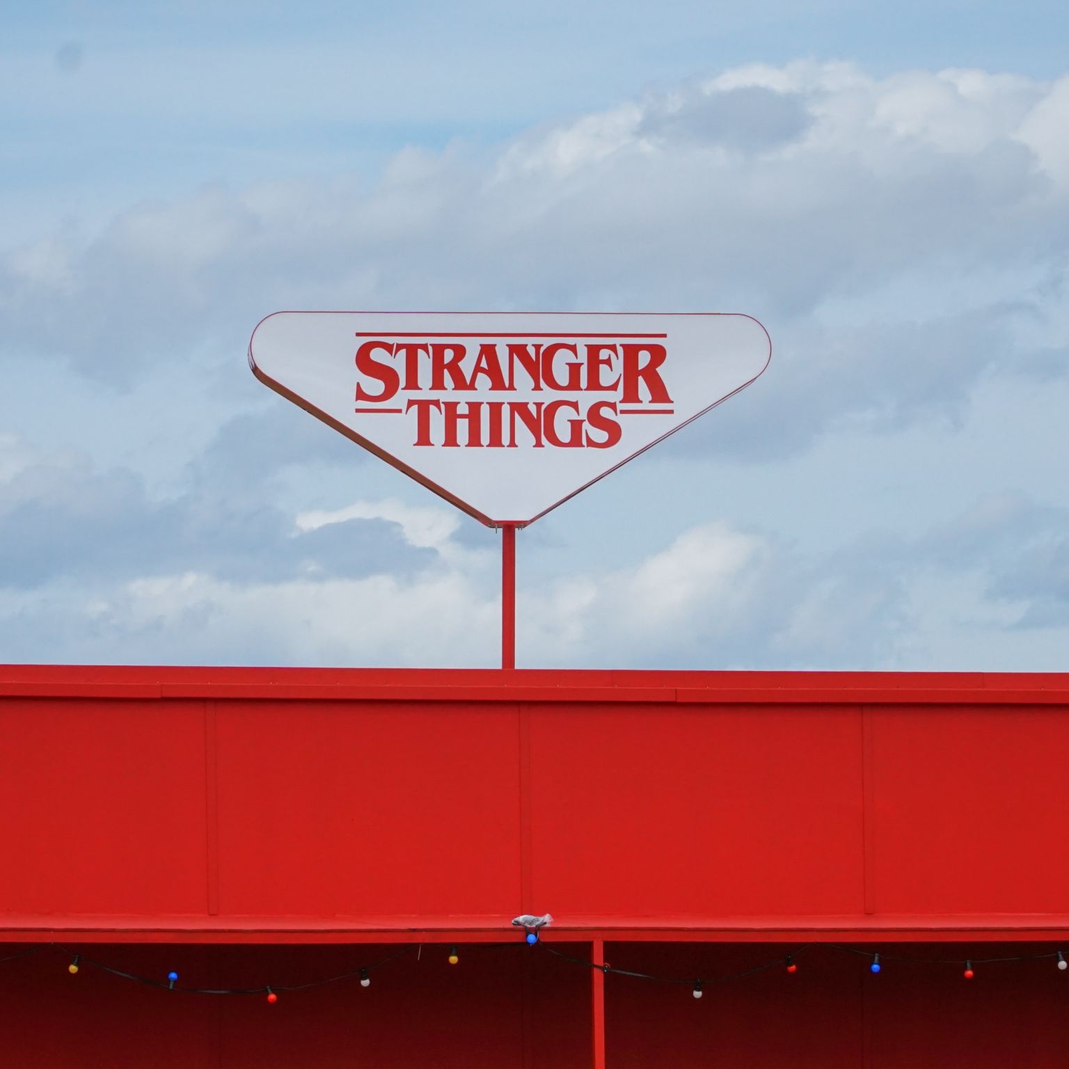 A triangular Stranger Things sign above a red building. There are clouds in the background behind the building.