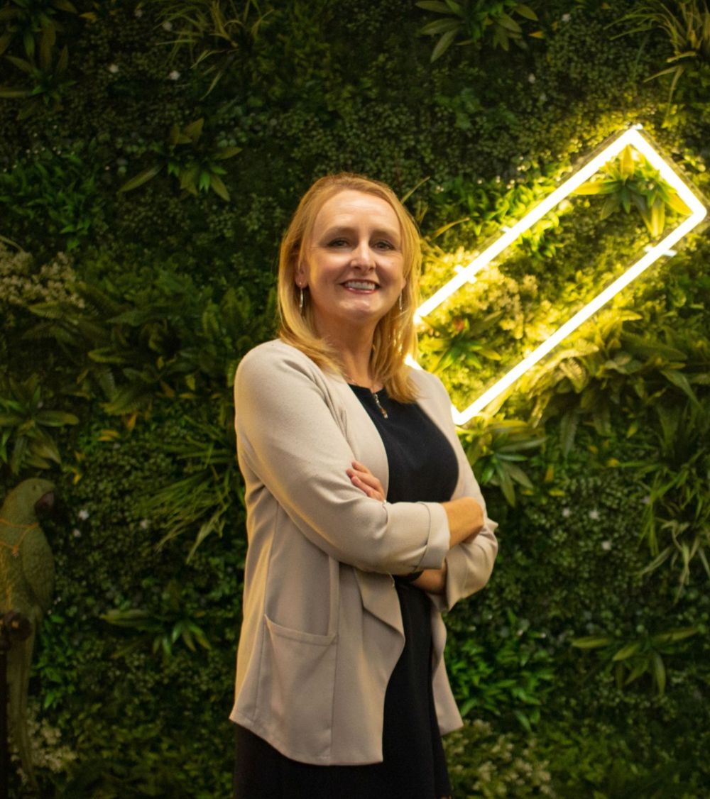 Gen Lilley-Walsh standing in front of a green plant wall with the Paradise logo glowing in the background. Gen has her arms folded and is beaming at the camera.