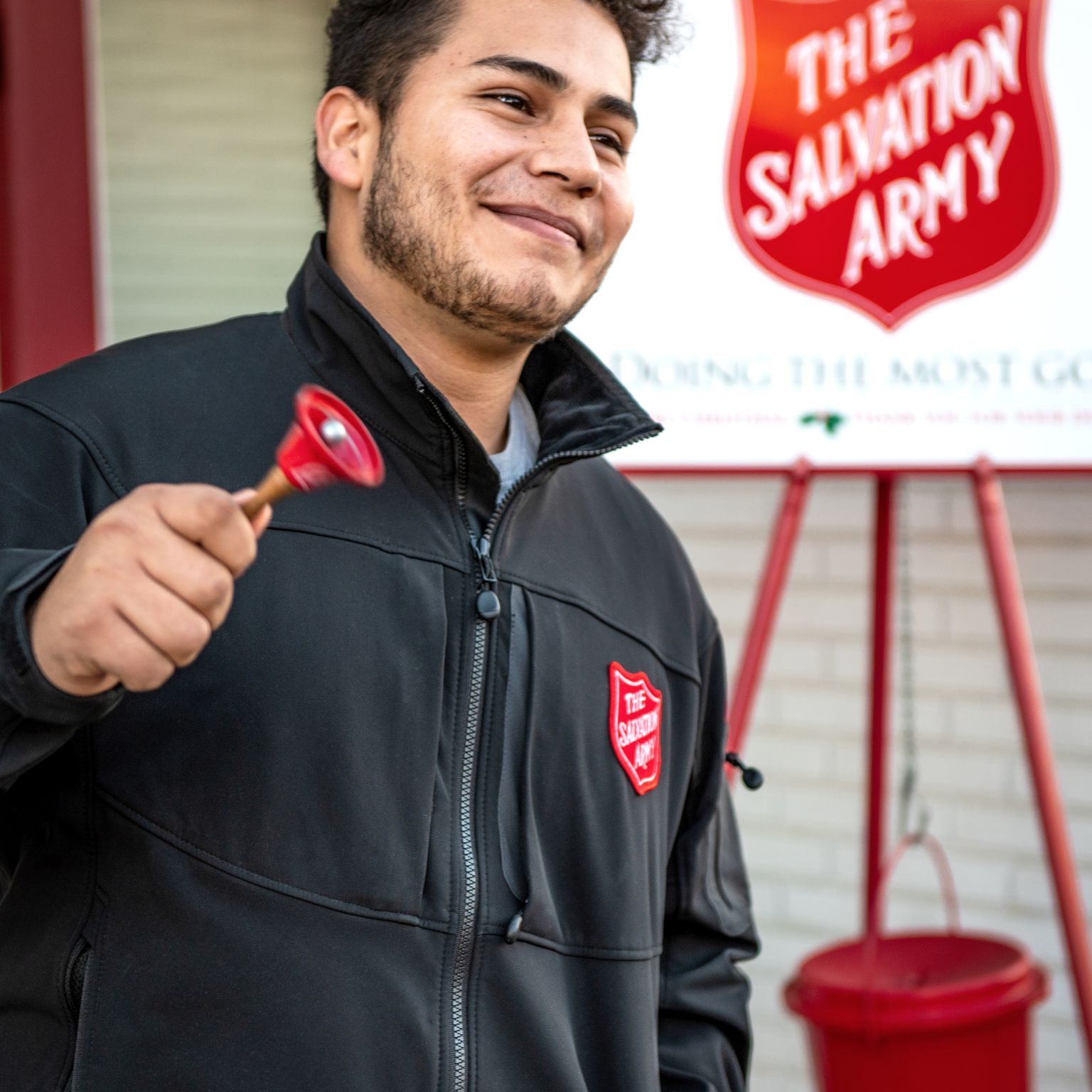 A young latino looking man rings a bell smiling, with a Salvation Army placard in the background.